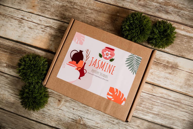 May include: A brown cardboard box with a sticker featuring pottery illustrations and the text "Jasmine Pottery Studios." The box is on a wooden surface, with two green decorative balls. The sticker has a pink background with orange and green accents.