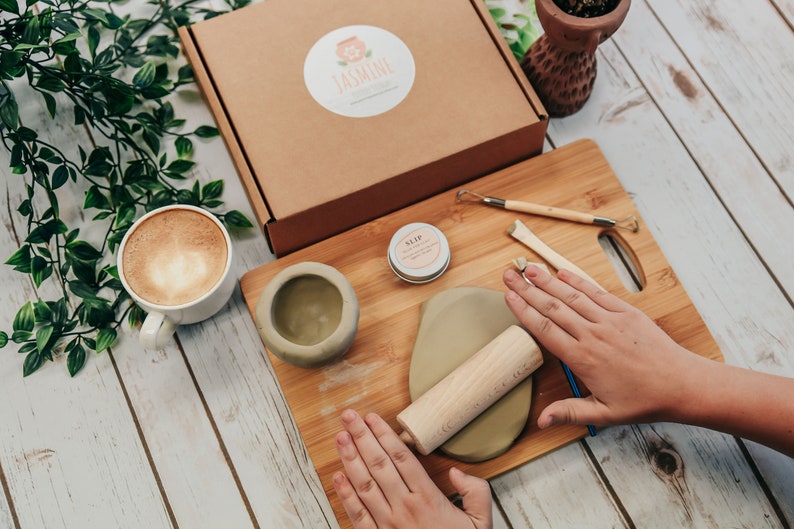 May include: A pottery kit with a wooden rolling pin, a small bowl, a container of slip, and two sculpting tools on a bamboo cutting board. The kit is on a white wooden table with a cup of coffee and a potted plant.