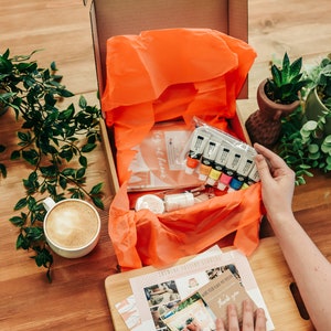 May include: An open box filled with art supplies, including paint tubes, brushes, and a canvas. The box is surrounded by greenery and a cup of coffee.