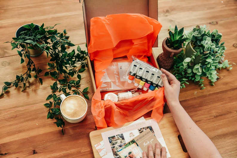 May include: An open box filled with art supplies, including paint tubes, brushes, and a canvas. The box is surrounded by greenery and a cup of coffee.