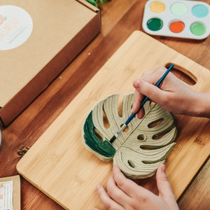 May include: A person paints a green monstera leaf-shaped ceramic coaster with a paintbrush. The coaster is on a wooden cutting board.