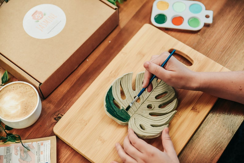 May include: A person paints a green monstera leaf-shaped ceramic coaster with a paintbrush. The coaster is on a wooden cutting board.
