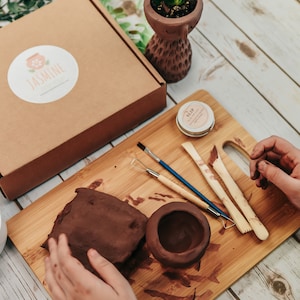 May include: A brown pottery kit with tools, clay, and a small container of slip. The kit is on a wooden cutting board with a hand reaching for the clay.
