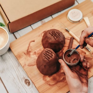 May include: A person is using a small brush to apply brown glaze to a clay pot. The pot has a textured surface and is sitting on a wooden cutting board. There is a cup of coffee and a box in the background.