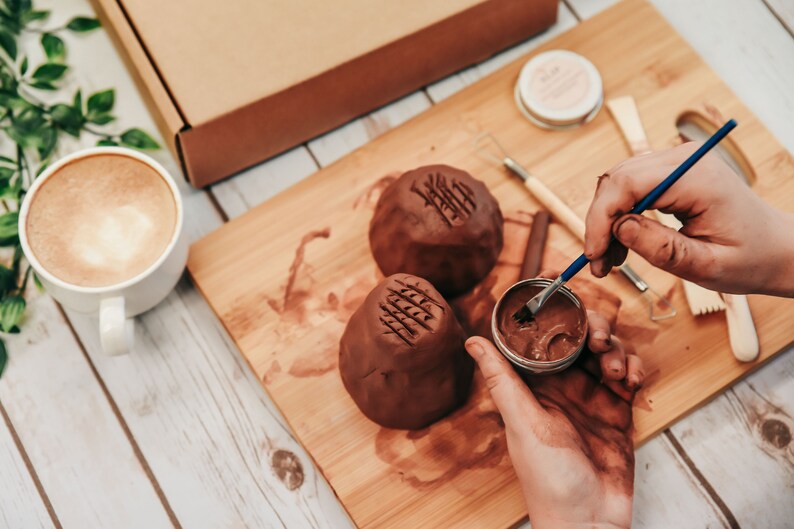 May include: A person is using a small brush to apply brown glaze to a clay pot. The pot has a textured surface and is sitting on a wooden cutting board. There is a cup of coffee and a box in the background.
