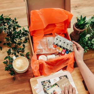 May include: An open cardboard box filled with art supplies, including paint tubes, brushes, and a small container. The box is lined with orange tissue paper. A cup of coffee and plants are on the wooden table.