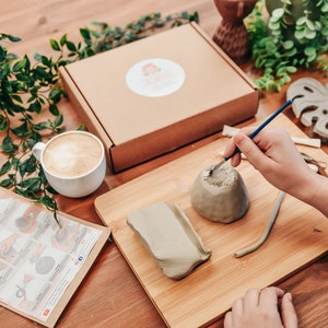 May include: A craft kit with clay, tools, and instructions for pottery. A person is using a brush to work on a small clay pot. A cup of coffee and a decorative leaf are also on the wooden table.