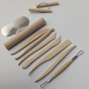 May include: A collection of light-colored wooden pottery tools, including a rolling pin, various shaping tools, and a wire tool. A metal blade and two wooden dowels with wire are also included. The tools are arranged on a white surface.