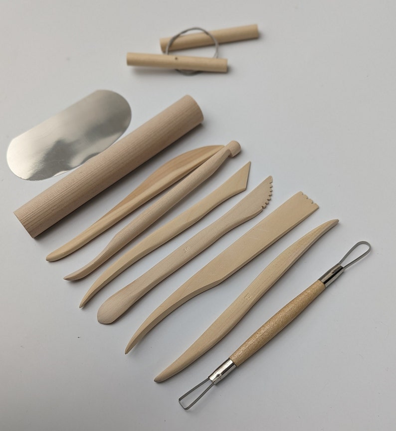 May include: A collection of light-colored wooden pottery tools, including a rolling pin, various shaping tools, and a wire tool. A metal blade and two wooden dowels with wire are also included. The tools are arranged on a white surface.