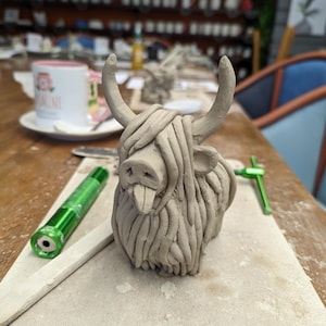 May include: A clay sculpture of a Highland cow with horns and shaggy fur. The sculpture is light brown and sits on a light-colored surface. A green tool and a green flashlight are nearby. A mug with text is in the background.