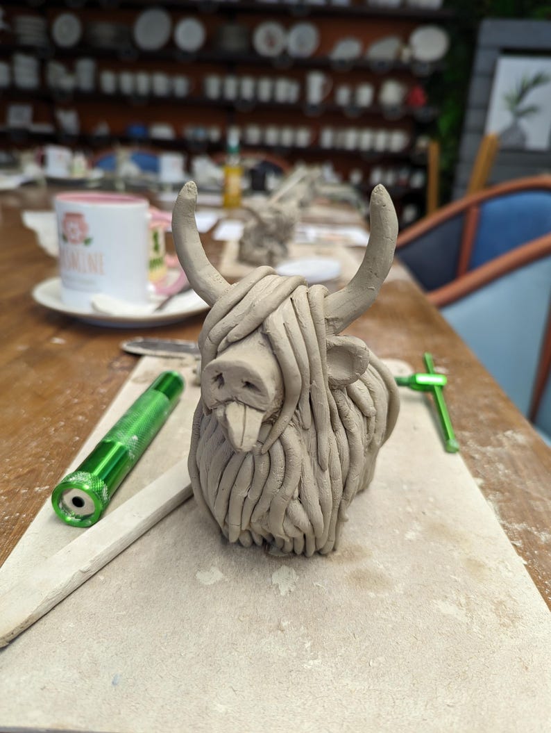 May include: A clay sculpture of a Highland cow with horns and shaggy fur. The sculpture is light brown and sits on a light-colored surface. A green tool and a green flashlight are nearby. A mug with text is in the background.