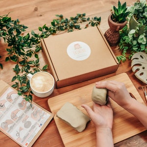 May include: A craft kit for making a small ceramic planter. The kit includes two blocks of air-dry clay, a small wooden cutting board, a set of sculpting tools, a small jar of sealant, and a set of instructions. The kit is on a wooden table with a cup of coffee and a few potted plants.