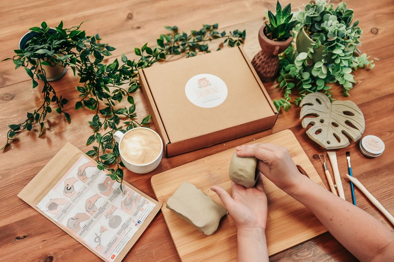 May include: A craft kit for making a small ceramic planter. The kit includes two blocks of air-dry clay, a small wooden cutting board, a set of sculpting tools, a small jar of sealant, and a set of instructions. The kit is on a wooden table with a cup of coffee and a few potted plants.