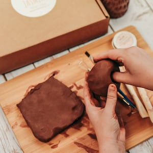 May include: A person is working with brown clay on a wooden cutting board. There is a block of clay and a ball of clay. The person is using their hands to shape the clay.