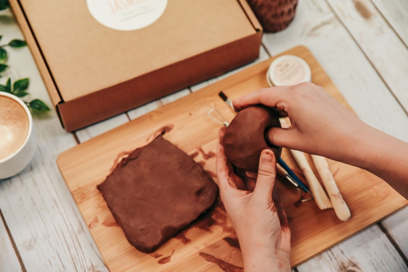 May include: A person is working with brown clay on a wooden cutting board. There is a block of clay and a ball of clay. The person is using their hands to shape the clay.