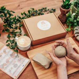 May include: A craft kit with clay, tools, and instructions. A ball of clay is held in the hands. A box with a logo, a cup of coffee, and decorative plants are also visible. The kit includes a wooden cutting board and a leaf-shaped item.