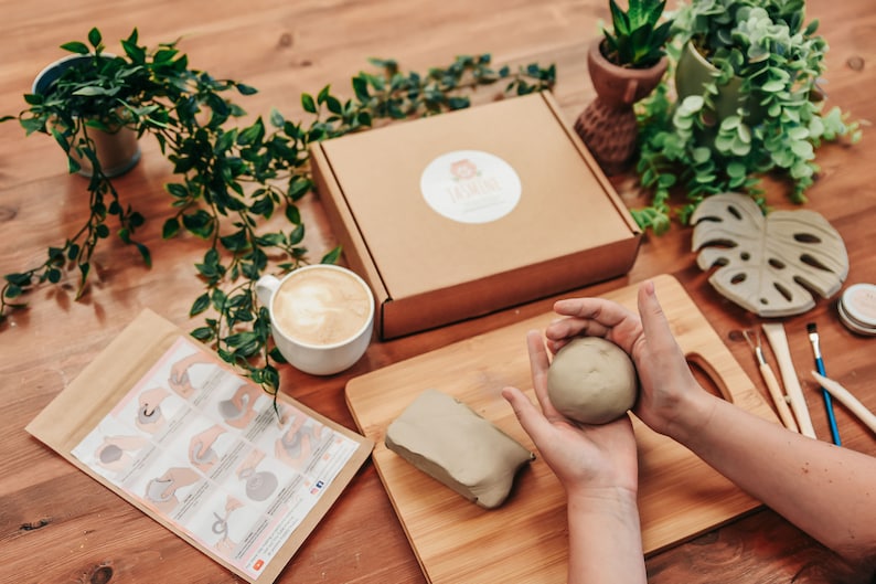 May include: A craft kit with clay, tools, and instructions. A ball of clay is held in the hands. A box with a logo, a cup of coffee, and decorative plants are also visible. The kit includes a wooden cutting board and a leaf-shaped item.