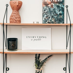 May include: A wooden shelf unit with three shelves, featuring a terracotta planter with a succulent plant, a black candle, a white framed print with the text "EVERYTHING is possible", and a book titled "POTTERS LIMITED EDITION".
