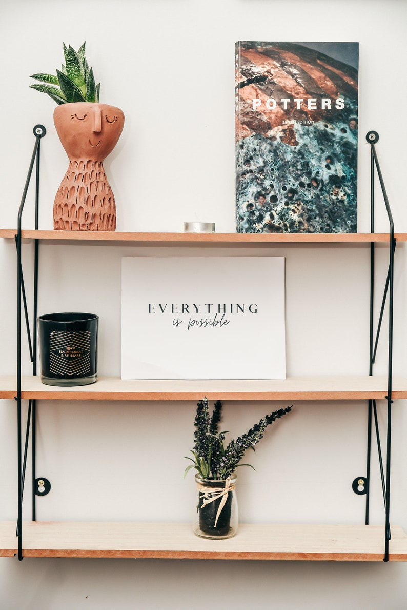 May include: A wooden shelf unit with three shelves, featuring a terracotta planter with a succulent plant, a black candle, a white framed print with the text "EVERYTHING is possible", and a book titled "POTTERS LIMITED EDITION".