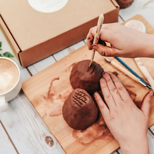 May include: A person using a wooden tool to create a design on a brown clay pot. The pot is sitting on a wooden cutting board with other tools nearby.
