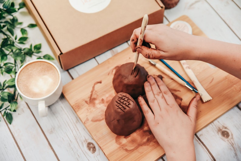 May include: A person using a wooden tool to create a design on a brown clay pot. The pot is sitting on a wooden cutting board with other tools nearby.