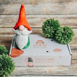 May include: A white box with the text "Jasmine Pottery Studios" and "Create a Gnome Air Dry Clay At Home Kit" on it. A red and white gnome figurine is sitting on top of the box. There are two green pom-poms on the wooden surface.