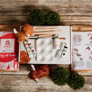 May include: A clay at home kit with various art supplies. The kit includes clay, paint tubes, a palette, brushes, and sculpting tools. The kit is packaged in a box with the text "Jasmine Clay at home". The kit also includes instructions.