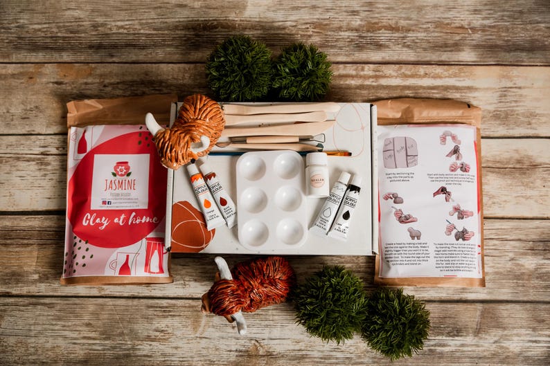 May include: A clay at home kit with various art supplies. The kit includes clay, paint tubes, a palette, brushes, and sculpting tools. The kit is packaged in a box with the text "Jasmine Clay at home". The kit also includes instructions.