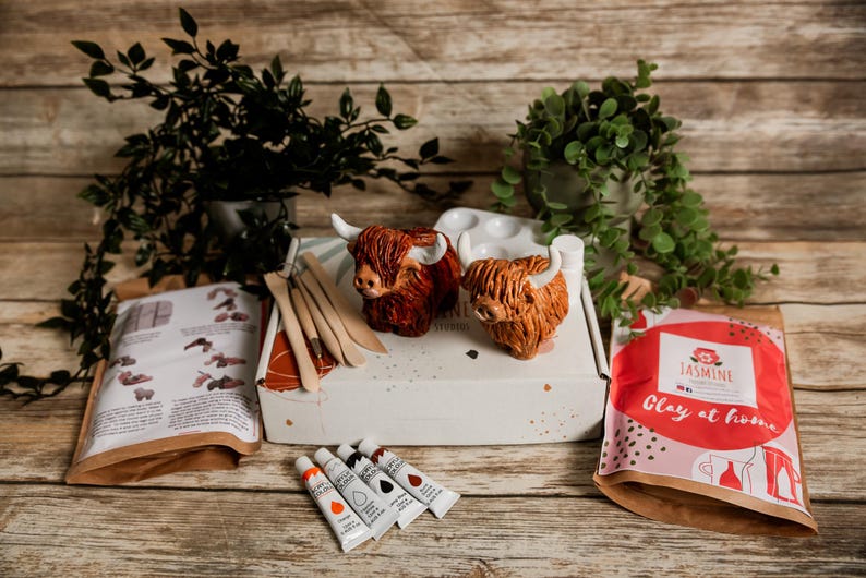 May include: A craft kit with two Highland cow sculptures, paint tubes, and tools. The kit includes a box with the text "Jasmine Studio" and a bag with the text "Clay at home". The sculptures are brown and orange.