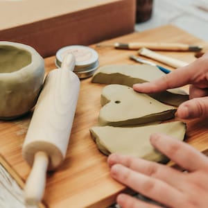 May include: A wooden rolling pin, a small bowl of clay, and several pieces of clay shaped like hearts are on a wooden cutting board. There are also several small tools for working with clay.