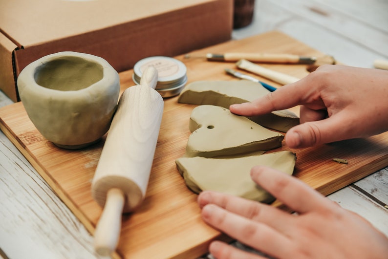 May include: A wooden rolling pin, a small bowl of clay, and several pieces of clay shaped like hearts are on a wooden cutting board. There are also several small tools for working with clay.