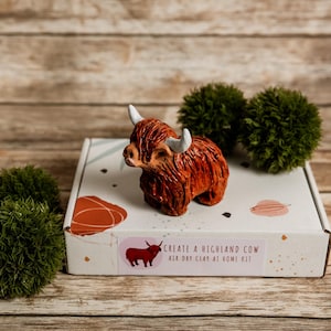 Puede incluir: Un kit de arcilla de vaca de las Tierras Altas con una figura marrón y blanca, una caja con el texto "Create a Highland Cow Air Dry Clay at Home Kit" y dos bolas decorativas verdes. La figura está sobre una caja blanca.