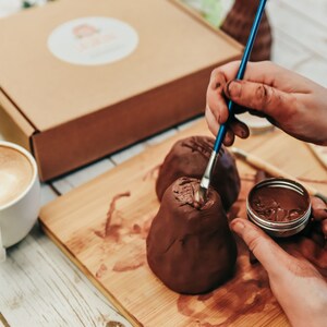 May include: A person paints a brown clay ornament with a blue paintbrush. The ornament is on a wooden surface with a small container of brown paint.