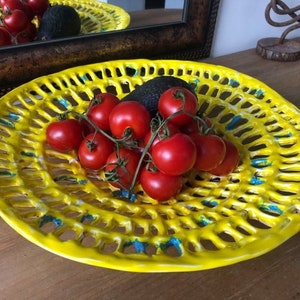 Op de afbeelding: Een gele keramische serveerschaal met een traliewerkontwerp en blauwe accenten. De schaal is gevuld met rode tomaten en een groene avocado.