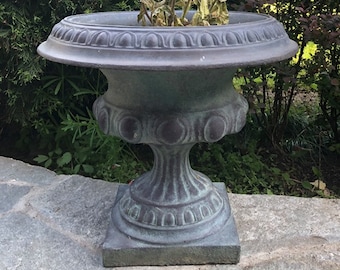 May include: A weathered, gray urn-shaped planter with a decorative rim and base, filled with vibrant purple flowers. The planter sits on a stone surface, surrounded by lush greenery, creating a classic garden aesthetic.