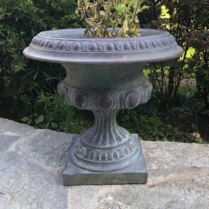 Puede incluir: Una jardinera en forma de urna grisácea y desgastada, con borde y base decorativos, llena de vibrantes flores moradas. La jardinera se asienta sobre una superficie de piedra, rodeada de exuberante vegetación, creando una estética de jardín clásica.
