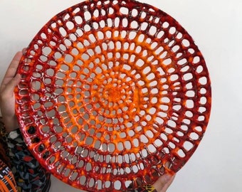 May include: A large, round, orange and red ceramic bowl with a circular, openwork design. The bowl is glazed and has a shiny finish.