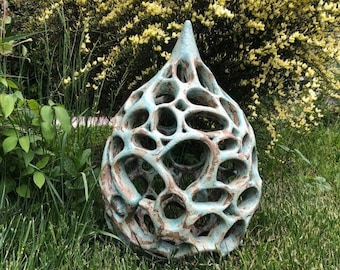 May include: A teal blue ceramic sculpture in the shape of a teardrop with a lattice design. The sculpture is sitting on a bed of green grass.
