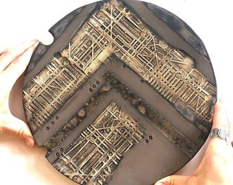 May include: A round ceramic plate with a black background and a gold geometric design. The design features a chevron pattern with a grid-like texture.