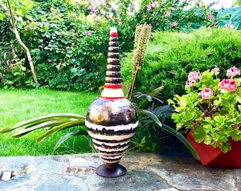May include: A decorative ceramic vase with a bulbous body and a tall, conical neck. The vase features a brown, black, and white striped pattern, with red accents near the top. It stands on a dark brown base, set against a backdrop of greenery.