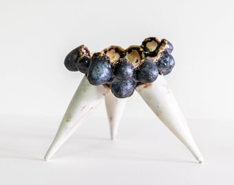 Op de afbeelding: Een keramische plantenpot met een zwarte en gouden gespikkelde afwerking. De pot heeft een afgeronde bovenkant en drie puntige poten.