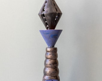Op de afbeelding: Een keramisch beeld met een geometrische, ruitvormige bovenkant, een kegelvormig middenstuk in blauw en paars en een meerlaags voetstuk in goud en blauw.