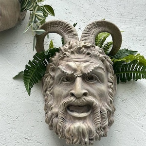 Puede incluir: Una escultura de piedra gris de la cara de un hombre con cuernos, barba y boca abierta. La escultura está montada en una pared blanca y tiene plantas verdes creciendo alrededor.