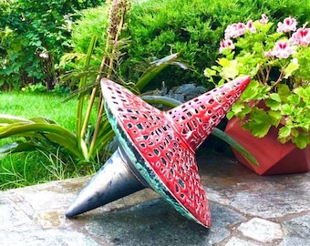 May include: A red ceramic spinning top with a black metal point. The top has a decorative, lacy design and is sitting on a stone patio.