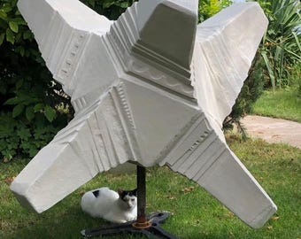 May include: A large, white concrete star-shaped garden sculpture with a black metal base. The sculpture is placed on a grassy lawn, and a black and white cat is resting beneath it.