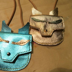 Puede incluir: Dos pequeñas carteras de cuero con caras de zorro. Una cartera es dorada con ojos verdes y la otra es azul con ojos dorados.