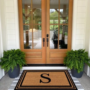 May include: A brown coir doormat with black trim and the letter "S" in black. The doormat is placed in front of a wooden double door with glass panels.