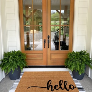 May include: A brown coir doormat with white and black striped edging. The doormat has the word "hello" written in black cursive script.