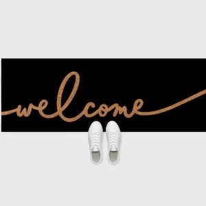 May include: A black rectangular doormat with the word "welcome" in a cursive, light brown font. Two pairs of white sneakers are at the bottom of the mat. The mat is on a white background.