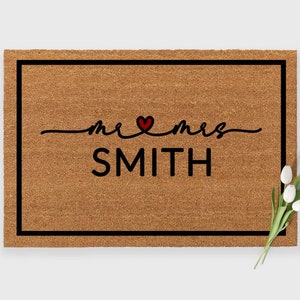 May include: A rectangular doormat with a black border and the words "mr & mrs SMITH" in black script, with a red heart symbol. The mat is made of a natural brown material. A small bouquet of white flowers is placed on the right side.